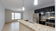 This single-floor, 2-bedroom, 2-bathroom residence offers a thoughtfully designed open-concept layout that maximizes functionality and flow. The kitchen serves as a central highlight, featuring granite countertops with subtle beige and gray veining, dark espresso cabinetry, and stainless steel appliances including a full-sized refrigerator. A kitchen island with an undermount sink and pendant lighting above creates a natural gathering point and provides additional workspace. The open layout connects the kitchen seamlessly to the living area, which benefits from a window that allows natural light to fill the space. The clean white walls and light flooring throughout contribute to a bright, contemporary ambiance. Modern recessed and pendant lighting fixtures ensure consistent illumination across the living and kitchen areas. The overall design aesthetic is sleek and minimalist, with dark cabinetry contrasting against light countertops and neutral walls. The efficient use of space throughout the unit makes it well-suited for comfortable everyday living while maintaining a polished, modern appearance.
