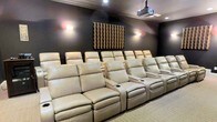 This single-floor home theater is purpose-built for an immersive cinematic experience, combining professional-grade design elements with refined comfort. The room is finished in deep, dark wall tones that minimize light reflection and enhance on-screen contrast, creating an atmosphere reminiscent of a commercial movie theater.

Seating is arranged in two tiered rows of plush cream leather recliners, each equipped with built-in cup holders and wide armrests, ensuring comfort during extended viewing sessions. The tiered layout ensures unobstructed sightlines from every seat in the room.

A ceiling-mounted projector serves as the primary display source, positioned to deliver a large, centered image onto the front screen. Acoustic treatment is addressed through a structured wood diffuser panel mounted prominently on the rear wall, helping to manage sound reflections and enhance audio clarity throughout the space.

Ambient wall sconces provide soft, warm accent lighting along the side walls, offering practical illumination without compromising the darkened viewing environment. Recessed ceiling lights supplement the lighting scheme. The overall design prioritizes acoustics, visual performance, and comfort, resulting in a polished and highly functional private screening room.