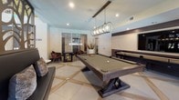 This thoughtfully designed single-floor amenity space makes a lasting impression from the moment you step through its elegant double-glass entry doors, flanked by sculptural tree-branch metalwork panels and built-in bench seating that set a nature-inspired tone throughout.

The main lounge is a showstopper, boasting soaring double-height ceilings adorned with a striking driftwood chandelier. Floor-to-ceiling windows and glass sliding doors flood the room with natural light and offer views of the surrounding outdoor grounds. A sleek, large-scale fireplace with a metallic surround anchors one wall, while plush seating — including a tufted ochre sofa and accent chairs — rests atop a layered area rug, creating a warm and inviting atmosphere.

Adjacent to the lounge, a well-appointed kitchenette area features rich wood cabinetry, marble-veined countertops, a coffee and beverage station, stainless steel appliances, and a bold floor-to-ceiling forest mural that brings an immersive, nature-inspired energy to the space.

The game room offers a more energetic atmosphere, complete with a substantial billiards table illuminated by a chic brass pendant light fixture, a shuffleboard table, arcade game, and comfortable lounge seating — all finished with large-format tile flooring and recessed lighting overhead.