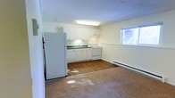 This single-floor space features an open-concept layout that combines the kitchen and living area into one cohesive, functional zone. The kitchen is outfitted with white cabinetry, a white refrigerator, a range, a dishwasher, and a sink set beneath upper and lower cabinets, providing ample storage. A fluorescent overhead light fixture illuminates the kitchen area, complementing the natural light that enters through a window on the adjacent wall. The flooring transitions from hardwood in the kitchen zone to carpet in the living area, visually distinguishing the two functional spaces while maintaining an open flow between them. A baseboard heater runs along the far wall, offering an efficient heating solution throughout the space. The walls are finished in a neutral off-white tone, contributing to a clean and bright atmosphere. The overall layout is practical and straightforward, making efficient use of the available footprint while keeping the space feeling open and uncluttered.