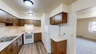 This single-floor unit offers a practical and well-organized layout across several distinct living areas. The kitchen is a standout feature, equipped with rich warm-toned wood cabinetry lining both upper and lower sections, white appliances including an electric range and full-size refrigerator, light countertops, and durable wood-look vinyl flooring. The layout provides ample storage and workspace for everyday use.

The living area welcomes residents through a wood-trimmed entry door, transitioning from hard flooring at the entryway to plush wall-to-wall carpeting that extends throughout the main living and sleeping spaces. The carpeted rooms are bright and neutral, with windows fitted with vertical blinds or standard frames that allow natural light to filter in, with views of the surrounding neighborhood.

The bedrooms are generously sized, each featuring carpet flooring, neutral walls, and windows that bring in daylight. One bedroom includes a large sliding wood-panel closet, offering substantial built-in storage. A wall-mounted air conditioning unit is also present in one of the rooms.

The full bathroom is well-appointed with wood-toned vanity cabinetry, a wide mirror, light countertops, wood-look vinyl flooring, and a full tub-shower combination. The overall ambiance of the space is clean, functional, and move-in ready.