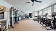 Located at 4490 Marietta Street in Powder Springs, GA, this fitness center offers a generously sized, single-floor layout designed to accommodate a comprehensive range of workout needs. The space features a clean, modern aesthetic with neutral-toned carpeted flooring, white walls, and recessed lighting throughout, creating a bright and inviting atmosphere. Large windows along one side of the room allow ample natural light to fill the space, while ceiling fans provide added airflow and comfort during workouts.

The cardio zone is well-appointed with multiple treadmills, elliptical machines, and stationary bikes, all arranged to allow easy access and movement between equipment. Adjacent to this area, the strength training section houses a Matrix cable machine system, weight benches, and a full dumbbell rack with an accompanying wall-mounted mirror. A dedicated functional training area toward the rear of the space features a multi-tiered equipment storage rack stocked with medicine balls, kettlebells, and resistance bands, set against a bold black accent wall with a wall-mounted flat-screen television. A spin bike rounds out the functional zone, making this fitness center a versatile facility suited for a wide variety of training styles and fitness goals.