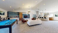 Located at 505 W Baseline Rd in Tempe, Arizona, this single-story community clubhouse offers a thoughtfully designed gathering space with a range of amenities for socializing and recreation. The open-plan layout features soaring vaulted ceilings and a neutral palette of whites and grays, accented by signature teal blue doors that lend a cohesive, contemporary character throughout.

The central lounge area is anchored by a plush white sectional sofa, cognac leather armchairs, and a custom built-in entertainment wall with teal-painted recessed shelving, warm wood cabinetry, and a flat-screen TV. A statement chandelier hangs overhead, complementing recessed lighting throughout. Adjacent to the lounge, a billiards table sits beside large windows that frame views of the surrounding palm-tree-lined grounds, flooding the space with natural light.

A stone-clad fireplace with a wood mantle serves as a dramatic focal point in the transitional area, paired with a floating wood media console. The kitchen is fully equipped with stainless steel appliances, a tile backsplash, stone countertops, and ample cabinetry, supported by a bar-height counter with gold-accented stools ideal for casual dining. A dedicated dining area accommodates a large communal wood table beneath a candle-style rectangular chandelier, with windows overlooking the outdoor pool area.