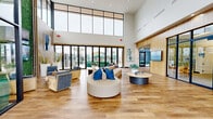 The Calm Luxury Living at Rainbow leasing and amenity center is a thoughtfully designed single-floor space spanning a generous footprint that immediately impresses with its dramatic double-height ceilings and expansive wall-to-wall clerestory windows. Natural light floods the open lobby and lounge area, which features warm hardwood-style flooring, a curved sectional sofa with vibrant blue accent pillows, and rounded coffee tables that create inviting conversation zones. A striking living green wall serves as a bold focal point, complemented by a slatted wood accent wall with mounted screens and curated artwork. The reception area near the entrance is finished with natural wood cabinetry and branded signage, offering a welcoming, professional first impression. Large multi-panel glass folding doors open directly to an outdoor courtyard, blurring the boundary between interior and exterior spaces. A dedicated E.Lounge provides a glass-enclosed business center with workstations and a tropical mural backdrop. A casual dining and refreshment area features pendant lighting, a coffee station, wine cooler, and a mounted display, offering residents a comfortable space to gather. Throughout, the design palette of warm wood tones, white walls, and blue accents creates a cohesive, resort-inspired ambiance.