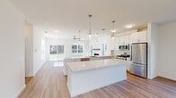Located at 20 Maple Lane in Farmingville, NY, this two-story home offers five bedrooms and four bathrooms across a well-designed layout. The main floor is the larger of the two levels, anchored by a bright, open-concept kitchen and living area. The kitchen is a standout feature, showcasing white shaker cabinetry, a large center island with quartz countertops, sleek pendant lighting, subway tile backsplash, and stainless steel appliances including a gas range and French door refrigerator. Light hardwood flooring flows throughout the main level, and large sliding glass doors and windows flood the space with natural light and offer outdoor views. A carpeted staircase with a wood handrail leads to the upper level, which hosts additional bedrooms and a hallway with hardwood-style flooring. The generously sized primary bedroom features plush carpet, a ceiling fan, and abundant natural light, accompanied by a walk-in closet with wire shelving. The primary bathroom is appointed with a dual-sink vanity, quartz countertops, white cabinetry, a large mirror, and a glass-enclosed shower. Additional bedrooms feature carpet, ceiling fans, and ample windows. A dedicated laundry room with a full washer and dryer rounds out the home's functional amenities.
