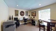 This single-floor business center is designed to provide a functional and comfortable workspace for productivity. The room features a built-in L-shaped desk that runs along two walls, offering generous counter space accommodating multiple computer workstations and printers, making it ideal for multi-tasking or simultaneous use by several individuals. Ergonomic black office chairs are positioned at each station, ensuring comfort during extended work sessions. A large window with blinds allows natural light to fill the room, creating a bright and welcoming atmosphere. The warm, neutral-toned walls are complemented by recessed ceiling lighting, which provides consistent, even illumination throughout the space. Decorative touches — including framed artwork, a distinctive sunburst mirror, and small potted plants — add character and warmth to what might otherwise be a purely utilitarian environment. A small seating area with a bistro-style table and chair is tucked near the window, offering an alternative spot for reading or casual work. The carpeted flooring contributes to a quiet, focused ambiance. Overall, this business center strikes a balance between professional functionality and an inviting, aesthetically considered interior.