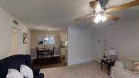 This single-floor residence offers a seamlessly connected layout that integrates the living, dining, and kitchen areas into one cohesive open-concept space. The living area is finished with plush wall-to-wall carpeting and features a ceiling fan with integrated lighting, providing both comfort and airflow. A front entry door with a sidelight panel opens directly into this welcoming space, accompanied by a small entryway accent table.

The dining area transitions naturally from the living space, showcasing hardwood-style flooring, a decorative area rug, and a classic pendant light fixture overhead. A dining table with chairs anchors the space, which is brightened by a window offering natural light. Adjacent to the dining area, the kitchen is equipped with white cabinetry, countertops, and a built-in microwave, creating a clean and functional cooking environment.

Throughout the space, neutral beige tones on the walls, a textured popcorn ceiling, and wainscoting-style trim details contribute to a traditional residential aesthetic. The open sightlines between rooms enhance the sense of space and connectivity, making this a practical and comfortable single-level home.