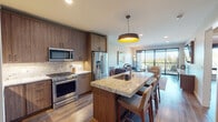 This thoughtfully designed single-floor residence offers a seamless blend of modern aesthetics and functional living. The open-concept kitchen is a true centerpiece, featuring rich walnut-toned cabinetry that reaches to the ceiling, a striking granite island with bar seating, a stone tile backsplash, and a full suite of stainless steel appliances including a gas range, built-in microwave, and French door refrigerator. A bold drum pendant light anchors the space, while warm wood-look flooring flows throughout and connects the kitchen to the living area. The living room is bright and inviting, benefiting from abundant natural light through expansive floor-to-ceiling sliding glass doors that open to an outdoor balcony with leafy views. The bedroom is a serene retreat with a wood-paneled accent wall, ceiling fan, large window framing outdoor greenery, and a wall-mounted television. The en-suite bathroom is spa-like, showcasing dual vanities with granite countertops, illuminated mirrors, and large-format tile. A dedicated hallway laundry area with stacked washer and dryer adds everyday convenience. Throughout, the space maintains a cohesive, contemporary palette of warm neutrals and natural wood tones.