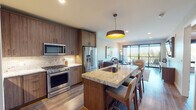 This thoughtfully designed single-floor residence offers a seamless blend of modern aesthetics and functional living. The open-concept kitchen is a true centerpiece, featuring rich walnut-toned cabinetry that reaches to the ceiling, a striking granite island with bar seating, a stone tile backsplash, and a full suite of stainless steel appliances including a gas range, built-in microwave, and French door refrigerator. A bold drum pendant light anchors the space, while warm wood-look flooring flows throughout and connects the kitchen to the living area. The living room is bright and inviting, benefiting from abundant natural light through expansive floor-to-ceiling sliding glass doors that open to an outdoor balcony with leafy views. The bedroom is a serene retreat with a wood-paneled accent wall, ceiling fan, large window framing outdoor greenery, and a wall-mounted television. The en-suite bathroom is spa-like, showcasing dual vanities with granite countertops, illuminated mirrors, and large-format tile. A dedicated hallway laundry area with stacked washer and dryer adds everyday convenience. Throughout, the space maintains a cohesive, contemporary palette of warm neutrals and natural wood tones.