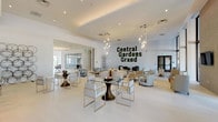 Central Gardens Grand is a elegantly designed single-story communal space that blends contemporary aesthetics with a welcoming, resort-like atmosphere. The expansive open floor plan is anchored by crisp white tile flooring and soaring ceilings, creating a bright and airy environment throughout.

The main lounge area is thoughtfully furnished with neutral-toned seating arrangements, including upholstered armchairs and glass-topped tables, arranged across patterned area rugs. Sculptural pendant light fixtures cascade from the ceiling, adding a striking decorative element overhead. A bold moss letter wall installation spelling "Central Gardens Grand" serves as the space's defining focal point, introducing a natural, organic texture against the clean white walls.

Floor-to-ceiling black-framed windows and glass doors line one side of the space, flooding the interior with natural light and providing direct views and access to an outdoor terrace area with surrounding greenery. A textured stone accent wall houses a wall-mounted flat-screen television, flanked by additional seating.

A well-appointed bar area features a long white countertop with woven detailing, lined with white upholstered barstools. Illuminated built-in shelving displays decorative objects alongside a television, while gold globe pendant lights add warmth and visual interest above.