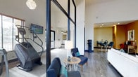 This single-floor space makes a strong architectural impression through its use of dramatic floor-to-ceiling glass partitions framed in bold black steel, which divide the open layout into distinct functional zones while maintaining a sense of openness and visual connectivity throughout.

The lounge area anchors the space with plush navy blue seating, a round accent table, and a stylish table lamp, creating a comfortable and polished gathering spot. Recessed lighting and soaring ceilings amplify the sense of volume, while large-format tile flooring in a cool grey tone runs seamlessly underfoot, lending a clean and cohesive aesthetic.

Adjacent to the lounge, a fitness area is visible through the glass partition, equipped with cardio equipment and a wall-mounted television — a practical and well-integrated feature of the overall layout. Further into the space, a bright reception or service counter area opens up, finished in crisp white tones that contrast with the bold accent colors used throughout.

To one side, a dining or bar area is partially visible, featuring high seating and a warm yellow accent wall that introduces a touch of color and warmth. Together, these zones combine to create a versatile, community-oriented space that balances modern design with functional amenities in a bright, well-appointed environment.