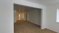 This single-story space presents a clean, functional layout characterized by open, flowing rooms connected through a wide arched passageway. The interior features wall-to-wall neutral-toned carpeting throughout, providing a uniform and cohesive aesthetic across the living areas. White painted walls and white trim contribute to a bright, airy atmosphere, while the wide coved archway that divides the main living space from an adjacent area adds a subtle architectural detail.

Looking through the passageway, the layout extends into what appears to be a hallway connecting additional rooms, including a door visible at the far end with a decorative screen or security door, allowing for natural light to filter into the interior. A second interior door is visible along the hallway, suggesting access to additional rooms or utility spaces. The lower ceiling in the inner section contrasts with the more open feel of the main room, creating a distinction between the spaces.

Overall, the space maintains a simple, no-frills design with clean lines and neutral finishes, making it a practical and adaptable environment well-suited for a variety of uses or furnishing styles.