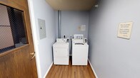 This single-floor space features a dedicated laundry room finished with light blue walls and warm wood-look flooring that together create a clean, functional environment. The room houses a side-by-side washer and dryer setup, both white Whirlpool appliances, accompanied by a coin-operated washing machine — suggesting shared or multi-unit use. A metal exhaust vent pipe runs vertically along the back wall, serving the dryer and venting upward through the ceiling. An electrical panel is mounted on the left wall, providing convenient access to the circuit breaker. Informational notices are posted near the appliances, offering instructions or usage guidelines for occupants. The space is compact yet efficiently organized, with white baseboards framing the perimeter and a standard wood door providing entry. The overall ambiance is utilitarian and tidy, designed to maximize functionality within a modest footprint.