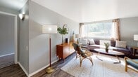 Located at 105 Honeysuckle Terr in Fairport, NY, this single-floor residence offers a well-organized layout with a cohesive, modern-casual aesthetic throughout. The living area is a welcoming space anchored by large windows that invite abundant natural light and offer pleasant outdoor views of the surrounding greenery. Rich wood-look flooring runs through the room, complementing the mid-century modern furnishing style, while a built-in closet provides convenient storage. The kitchen features warm wood cabinetry, light countertops, a gas range, and a single-basin sink, all set against soft gray walls and wood-look flooring — a practical and tidy space for everyday cooking. The bedroom is a restful retreat with plush carpeting, a large window framing natural light, and clean gray walls that create a calm, neutral atmosphere. A circular gold-framed mirror adds a stylish accent above the bed. The bathroom is finished with gray walls, wood-look flooring, a white vanity with a framed mirror, and a shower-tub combo with a classic curtain. Each area of this residence reflects a thoughtful, functional design that balances comfort and style in a compact, efficient footprint.