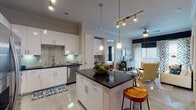Located at 6600 McKinney Ranch Pkwy in McKinney, Texas, this single-floor residence offers a thoughtfully designed layout with modern finishes and a cohesive contemporary aesthetic throughout. The kitchen is a standout feature, showcasing white shaker cabinetry, dark granite countertops, a tile backsplash, and a full suite of stainless steel appliances including a refrigerator, range, microwave, and dishwasher. A central island with bar seating and pendant lighting adds both function and style, while track lighting and crown molding elevate the space further. A built-in desk area provides a convenient workspace just off the kitchen. The open-concept living area is bright and inviting, featuring a ceiling fan, sliding glass doors leading to a private balcony, and bold patterned drapery that adds visual interest. The bedrooms offer natural light through large windows, crown molding, and ceiling fans, with one bedroom featuring a striking deep blue accent wall. The bathroom is well-equipped with a soaking tub, tiled shower surround, a dual-sink vanity with dark granite countertops, and direct access to a spacious walk-in closet with double hanging rods. Light wood-style flooring runs throughout the main living areas, contributing to the clean, modern ambiance of the home.