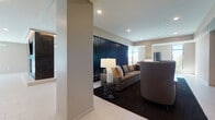 This single-floor communal space is designed with a contemporary aesthetic, combining functionality and style across several distinct zones. The main lounge area is expansive and light-filled, anchored by floor-to-ceiling windows that frame outdoor views and flood the interior with natural light. A large U-shaped sectional sofa in warm cognac tones sits atop a patterned area rug, complemented by a central bar-height communal table with sleek charcoal seating — ideal for both relaxed socializing and casual work. Recessed lighting and tray ceiling details add architectural depth throughout.

A secondary lounge area offers a more intimate setting, featuring neutral-toned sofas, a dark vertical slat accent wall, and layered lighting from table lamps and recessed fixtures. The kitchen is well-appointed with stainless steel appliances, wood-toned cabinetry, a dark tile backsplash, and a generously sized island with leather-and-gold bar stools beneath pendant track lighting — a striking centerpiece for the space.

The dining area accommodates a large group around a dark wood table, bathed in natural light from full-height sliding glass doors that open to a pool and courtyard area beyond. Throughout, large-format light tile flooring unifies the space, creating a clean, cohesive, and welcoming environment.