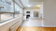 Located at 12-26 Rosseau Rd in Toronto's Bathurst Manor neighbourhood, this one-bedroom, one-bathroom unit offers a well-organized layout across a single floor. Warm parquet flooring runs throughout the living and bedroom areas, providing a classic aesthetic that contrasts nicely with the bright, neutral walls.

The kitchen is a standout feature, designed in a galley configuration with white shaker-style cabinetry extending to the ceiling for ample storage. Stainless steel appliances, including a refrigerator, stove, and microwave, complement light stone-effect countertops. A breakfast bar peninsula creates an open transition toward the living area, which benefits from large windows admitting generous natural light. A cast-iron radiator beneath the windows provides efficient heating.

The bedroom is a comfortable size, featuring two closets and a large window overlooking a green outdoor area and neighbouring buildings, offering a pleasant, open view. Parquet flooring continues here, maintaining visual consistency throughout.

The bathroom is clean and functional, finished with white subway-style wall tiles and a full bathtub with shower, accented by a small window providing natural ventilation. A connecting hallway ties all rooms together efficiently, completing this practical and well-presented unit.