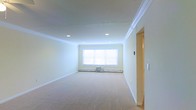 This well-maintained single-floor space exudes a clean, airy ambiance throughout. The main living area is finished with soft, plush carpeting underfoot and crisp white walls that reflect natural light, creating a bright and open feel. Elegant crown molding runs along the ceiling perimeter, adding a refined architectural detail that elevates the overall aesthetic. Recessed lighting is thoughtfully installed in the ceiling, providing consistent and even illumination across the room. A prominent window fitted with blinds serves as the focal point of the main room, allowing natural light to filter in and contributing to the space's welcoming atmosphere. A baseboard heating system runs beneath the window, along with a wall-mounted air conditioning unit, ensuring year-round comfort. The doorway leading to an adjacent room hints at additional functional areas within the layout. The overall design is simple, neutral, and move-in ready, making it highly versatile for a variety of uses. The combination of classic molding details, soft flooring, and ample lighting gives the space a timeless, comfortable character suited to everyday living.