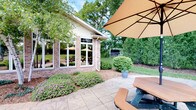 This single-story property showcases a classic brick exterior complemented by large, multi-pane white-framed windows that flood the interior with natural light while offering sweeping views of the surrounding landscape. The building's clean architectural lines and white trim details lend it a polished, well-maintained appearance.

Outside, a spacious stamped concrete patio serves as a welcoming outdoor gathering area, furnished with a picnic-style table and chairs shaded by a large tan market umbrella. The patio is thoughtfully bordered by decorative landscaping, including neatly trimmed flowering shrubs with soft pink blooms and rounded ornamental bushes set against a mulched garden bed.

The expansive backyard beyond the patio is lush with mature trees and well-kept green lawn, creating a serene, park-like setting. A chain-link fence along the perimeter provides definition to the outdoor space, which feels both open and private thanks to the dense tree canopy. Potted plants add additional touches of greenery throughout the patio area.

The combination of durable brick construction, generous window placement, and beautifully maintained outdoor spaces makes this property an inviting and functional environment, well-suited for both relaxation and social gatherings in a tranquil, nature-surrounded setting.
