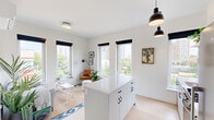 This single-floor residence showcases a clean, contemporary design centered around an all-white palette and light wood flooring throughout. The open-plan kitchen and living area forms the heart of the space, where a white kitchen island with black ring-pull hardware anchors the room beneath striking matte black pendant lights. Multiple windows dressed with bold black roller shades flood the area with natural light and frame views of surrounding greenery and the urban skyline beyond.

The living area flows seamlessly from the kitchen, offering a comfortable, well-lit space with city views visible through large windows. A dedicated sitting nook with corner windows on two sides creates an especially bright and inviting retreat.

Both bedrooms are quiet and well-appointed, each featuring windows with outdoor views, light wood flooring, and wall-mounted mini-split climate control units for individual temperature management. One bedroom includes a walk-in closet with built-in hanging storage, while the hallway also features a louvered-door closet for additional organization.

The bathroom presents a polished, minimal aesthetic with a white vanity, white countertop, large mirror, and a distinctive stained concrete floor that adds warmth and texture. The overall ambiance of the space is bright, calm, and modern throughout.