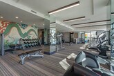Located at 811 Juniper St NE in Atlanta's Midtown neighborhood, this well-appointed fitness center offers a comprehensive and visually dynamic workout environment. The main exercise floor is generously sized, featuring multicolored striped carpet flooring and a clean, modern aesthetic with recessed and linear pendant lighting set against a white ceiling with architectural beam details. A striking hand-painted dragon mural anchors one end of the space, adding an artistic focal point that sets this gym apart. The main floor is equipped with a full array of cardio machines — including treadmills, stair climbers, and ellipticals — as well as a dedicated free weights area with a complete dumbbell rack and weight benches, all reflected in floor-to-ceiling mirrors. Floor-to-ceiling windows along one wall flood the cardio zone with abundant natural light and offer open views of the surrounding Atlanta cityscape and treeline. A separate spin studio, enclosed by glass partition walls, houses stationary cycling bikes alongside a wall-mounted display screen, bathed in soft natural light through sheer floor-length curtains. The entry area features a built-in counter station, a water fountain, and posted fitness center guidelines, ensuring a well-organized and user-friendly experience. The overall ambiance is energetic yet refined, blending contemporary design with functional amenities.