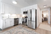 Located at 2613 Atlantic Ave in Virginia Beach, Virginia, this single-floor coastal retreat offers a thoughtfully designed layout with a bright, welcoming ambiance throughout. Light hardwood flooring runs continuously through the main living areas, tying the space together with warmth and cohesion.

The kitchen is well-appointed with crisp white shaker cabinetry, white quartz countertops, and a herringbone tile backsplash that adds a refined textural detail. Stainless steel appliances — including a range, over-the-range microwave, and dishwasher — are complemented by under-cabinet lighting that enhances both function and atmosphere.

The living area is bright and airy, featuring large sliding glass doors that open to a balcony with outdoor views, allowing natural light to flood the space. Sheer curtains frame the doors, contributing to the relaxed, coastal feel of the room. A wall-mounted television and ample seating make this an ideal space for both relaxing and entertaining.

The bedroom is a calm, neutral retreat with light hardwood floors, a substantial wood-frame bed, and soft, beach-inspired décor. Flanking nightstands and decorative wall panels above the headboard complete the serene aesthetic.

The bathroom features classic black-and-white mosaic penny tile flooring, a fully enclosed glass walk-in shower with white subway tile surround, and clean white fixtures — a timeless combination that suits the overall coastal character of the space.