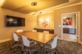 This single-floor conference room is a thoughtfully designed meeting space that balances functionality with contemporary style. At its center sits a substantial walnut conference table surrounded by sleek white chairs with chrome bases, comfortably accommodating up to six attendees. The room's distinctive ceiling features a raised tray detail finished in a warm gold tone, anchoring a modern circular pendant light fixture that serves as the space's focal point and provides ambient illumination. A large wall-mounted flat-screen display is positioned prominently for presentations and collaborative work. Patterned carpet in deep earth tones adds texture and visual interest underfoot. Natural light filters in through two windows fitted with solar shades, offering views of the surrounding urban environment. A dark-cabinetry credenza lines one wall, providing additional storage and surface space, while curated pop-art style prints add a vibrant, creative character to the room. One wall consists largely of floor-to-ceiling glass panels, offering visual connectivity to an adjacent lounge and amenity area, enhancing the sense of openness. A private entry door provides separation when needed. Overall, the space presents a professional yet inviting atmosphere well-suited for meetings, presentations, and focused collaboration.