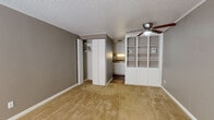 This single-floor studio offers a well-organized, open-concept layout that efficiently combines living, sleeping, and dining areas into one cohesive space. The main living area is finished with warm, neutral-toned carpet and painted in calming gray tones throughout, creating a clean and understated aesthetic. A ceiling fan overhead provides both comfort and a subtle design accent, while a textured ceiling adds a classic touch.

A standout feature of the main area is the built-in white shelving unit, providing ample display and storage space. Adjacent to it, a set of white closet doors offers practical wardrobe storage, keeping the living space uncluttered. A window near the entry door allows natural light to filter in, brightening the space alongside a wall-mounted HVAC unit for year-round climate control.

The kitchen is compact yet fully equipped, featuring white cabinetry, dark granite countertops, a coil-top range, a stainless steel sink, and under-cabinet lighting that lends a warm glow to the workspace. Dark hardwood flooring in the kitchen and bathroom areas provides a rich contrast to the carpeted living space.

The bathroom is neatly appointed with a white vanity, hardwood-style flooring, and a clean, straightforward layout that includes a toilet and towel fixtures. Overall, this studio presents a functional and cohesive living environment with practical storage and comfortable finishes throughout.