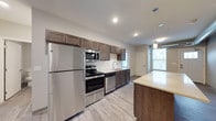 This single-floor space showcases a thoughtfully designed, modern kitchen as its centerpiece. The kitchen features a full suite of stainless steel appliances, including a refrigerator, electric range, built-in microwave, and dishwasher, all seamlessly integrated into a sleek, functional layout. Medium-toned wood cabinetry lines the walls from counter to ceiling, providing generous storage, while a white subway tile backsplash adds a clean, contemporary contrast. The light-toned countertops extend to an island or peninsula, offering additional workspace and casual dining potential. Pendant lighting hangs above this surface, complementing the recessed ceiling lights that illuminate the space evenly. Light wood-toned flooring runs throughout, contributing to a warm yet modern aesthetic. A window above the sink introduces natural light into the kitchen, while a larger window visible in the background floods the adjacent area with additional daylight. The open-concept design allows the kitchen to flow naturally into the surrounding living space, creating a cohesive and practical environment. The overall ambiance is clean, contemporary, and move-in ready.