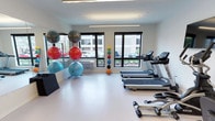 This fitness center offers a clean, modern environment designed to support a well-rounded workout routine. The space is set on a single level and is finished with bright white walls and a smooth, light-toned flooring that creates an open and energizing atmosphere. Large black-framed windows line the walls, allowing ample natural light to fill the room and providing views of the surrounding outdoor area, contributing to an airy and welcoming feel. Overhead linear lighting supplements the natural light, ensuring the space is well-illuminated throughout. The cardio section features a row of treadmills positioned to take advantage of the window views, making for an engaging workout experience. A dedicated functional training area includes a rack of medicine balls in a range of sizes and weights, as well as a collection of stability and exercise balls stored on a freestanding rack. A full-length mirror is mounted along one wall, providing visual feedback during workouts and further enhancing the sense of space. The overall layout is thoughtfully organized to maximize usability while maintaining an uncluttered environment. This fitness center is well-suited for a variety of training styles, from cardio and endurance work to balance and functional fitness training.