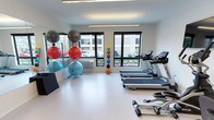 This fitness center offers a clean, modern environment designed to support a well-rounded workout routine. The space is set on a single level and is finished with bright white walls and a smooth, light-toned flooring that creates an open and energizing atmosphere. Large black-framed windows line the walls, allowing ample natural light to fill the room and providing views of the surrounding outdoor area, contributing to an airy and welcoming feel. Overhead linear lighting supplements the natural light, ensuring the space is well-illuminated throughout. The cardio section features a row of treadmills positioned to take advantage of the window views, making for an engaging workout experience. A dedicated functional training area includes a rack of medicine balls in a range of sizes and weights, as well as a collection of stability and exercise balls stored on a freestanding rack. A full-length mirror is mounted along one wall, providing visual feedback during workouts and further enhancing the sense of space. The overall layout is thoughtfully organized to maximize usability while maintaining an uncluttered environment. This fitness center is well-suited for a variety of training styles, from cardio and endurance work to balance and functional fitness training.