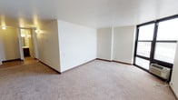 This single-floor space offers a clean, open layout designed for versatile living. The main living area is finished with neutral wall-to-wall carpeting that adds warmth underfoot, while the crisp white walls provide a bright, airy backdrop throughout. Dark wood baseboards offer a subtle contrast that frames each room with a classic, refined touch. A standout feature of the space is the large floor-to-ceiling window that floods the interior with abundant natural light and opens the room to exterior views, creating a sense of openness and connection to the outside. The layout includes a partial interior wall that defines distinct zones within the open-plan area, allowing for flexible furniture arrangements and functional use of the space. Recessed wall sconces provide additional ambient lighting. The overall ambiance is clean, minimal, and move-in ready, presenting a neutral canvas well-suited for a variety of design preferences. The straightforward single-level layout ensures ease of movement throughout, making the space both practical and comfortable for everyday use.
