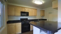 This two-story residence offers a practical and well-organized layout across its multiple levels. The kitchen serves as a functional centerpiece, featuring light maple-toned cabinetry with arched door detailing that provides generous storage space along the walls. Dark granite-style countertops contrast sharply with the lighter cabinetry, creating a bold visual dynamic throughout the space. Black appliances, including a gas range and built-in microwave overhead, complement the darker countertop palette cohesively. A peninsula extends into the kitchen area, adding additional counter space ideal for meal preparation or casual dining. The sink is positioned along the countertop run, offering a practical workspace. Flush ceiling lighting provides consistent illumination throughout the kitchen. Dark hardwood-style flooring grounds the space and ties together the contrasting elements of light cabinetry and dark surfaces. The overall aesthetic of the kitchen is clean and straightforward, combining classic cabinetry design with contemporary dark finishes. The two-floor layout suggests additional living, sleeping, and utility spaces spread across both levels of the home.