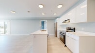 This single-floor unit offers a thoughtfully designed layout accommodating four bedrooms, one bathroom, and a dedicated laundry room. The open-concept kitchen and living area serves as the heart of the home, featuring a central island with an integrated sink, sleek white cabinetry with high-gloss upper cabinets, stainless steel appliances including an electric range and refrigerator, and a under-cabinet range hood. Light wood-tone flooring runs throughout, complementing the soft neutral wall tones that lend a bright, airy feel to every room. The living area benefits from large windows with slatted blinds and a sliding glass door that invites natural light and provides access to an outdoor space with open views. Each bedroom is generously proportioned, with the primary bedroom offering ample space, and the bedrooms feature sliding-door closets with built-in wire shelving for practical storage. The bathroom is finished in clean white with a full soaker tub and shower surround. A dedicated laundry room houses a stacked washer and dryer unit, adding everyday convenience. Recessed and flush-mount ceiling lighting throughout ensures consistent, well-distributed illumination. The overall ambiance is modern, clean, and functional, making this space well-suited for shared or family living.