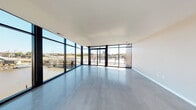 This contemporary waterfront unit offers a striking open-concept design defined by expansive floor-to-ceiling windows that frame sweeping views of a marina and surrounding waterway. The generous main living area is anchored by polished concrete flooring that reflects natural light throughout the space, creating a bright, airy atmosphere that flows seamlessly toward the glazed exterior walls and direct outdoor access via sliding glass doors.

The well-appointed kitchen is a standout feature, showcasing rich dark-stained cabinetry, a white subway tile backsplash, stainless steel appliances including a gas range and refrigerator, a stainless farmhouse sink, and a large center island topped with light quartz countertops. Pendant globe lights add a refined, modern touch overhead.

The bedrooms are carpeted for comfort, with at least one featuring sliding glass door access to an outdoor balcony and city views. Two full bathrooms serve the unit — a primary bath with a double-sink vanity featuring dark cabinetry, white countertops, a large mirror, and stylish lighting, and a secondary bath with a single vanity and similar contemporary finishes. Both bathrooms include accessibility grab bars. The overall design palette of neutral tones, warm wood accents, and industrial-modern details gives this space a sophisticated, urban character.