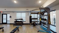 This modern fitness center is thoughtfully designed to accommodate a wide range of workout styles and fitness goals. The main cardio area is anchored by multiple treadmills, an elliptical, and a rowing machine, all positioned along a textured geometric accent wall with large windows that invite natural light and outdoor views of what appears to be a pool courtyard. A ceiling fan overhead adds to the comfort of the open, airy space.

A dedicated training zone provides a comprehensive strength and functional fitness experience, featuring a full rack of dumbbells, kettlebells, medicine balls, a TRX suspension system, battle ropes, and a heavy Everlast punching bag — all organized around a multi-functional training rack. An adjustable flat bench completes this well-stocked area.

Adjacent to the main floor, a glass-walled spin studio houses stationary cycling bikes and a wall-mounted TV, providing an immersive and semi-private environment for cycling workouts. A service counter with open shelving — stocked with towels and amenities — sits at the entrance, lending a polished, professional feel to the facility.

Throughout the space, light hardwood-style flooring, recessed lighting, and a clean neutral color palette with dark accent baseboards create a cohesive and energizing atmosphere. A water fountain and mechanical room are also conveniently located within the facility.