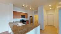 This single-floor residence centers around a functional and stylish kitchen that serves as the heart of the space. The kitchen is outfitted with warm medium-toned wood cabinetry providing ample storage, complemented by granite countertops with natural variation in tone. A peninsula-style counter extends outward, offering additional prep space and the potential for casual seating. Stainless steel appliances, including a refrigerator, gas range, and built-in microwave, lend a modern, cohesive look throughout the cooking area. A double basin sink is integrated into the counter, positioned conveniently near the range.

Track lighting runs along the ceiling, providing directed illumination throughout the kitchen. Light-toned tile flooring extends throughout the visible areas, contributing to a clean and cohesive aesthetic. The layout opens toward a hallway that leads to additional rooms, maintaining an easy flow through the space. Notably, a dedicated laundry area is visible just off the kitchen, housing a washer and dryer for added convenience. White walls and neutral finishes throughout create a bright, welcoming atmosphere.