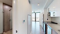 Located on Peachtree Street in Atlanta's vibrant Midtown neighborhood, this efficiently designed high-rise unit offers a thoughtful layout across a single floor. The open living area is anchored by dramatic floor-to-ceiling windows that flood the space with natural light and frame impressive panoramic views of the Atlanta skyline and surrounding cityscape. Hardwood flooring runs throughout the main living areas, adding warmth to the clean, modern aesthetic defined by crisp white walls and recessed lighting. The galley-style kitchen is well-appointed with stainless steel appliances, including a refrigerator and range, white cabinetry, and open shelving that maximizes storage in a compact footprint. A ceiling fan adds a functional touch to the airy living space. The bathroom features large-format tile flooring, a wood-accented vanity with an undermount sink, a wall-hung toilet, and a glass-enclosed walk-in shower with earthy brown tile surround. Smart home features are evident, including a digital thermostat and modern electrical controls throughout. The overall ambiance is contemporary and sleek, ideal for urban living in one of Atlanta's most dynamic corridors.