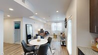 This single-floor residence features an open-concept layout that seamlessly connects the dining and living areas, creating a cohesive and functional living space. The interior is finished with light-toned hardwood-style flooring that runs throughout, contributing to a bright and airy atmosphere.

The dining area is anchored by a round marble-top table paired with charcoal upholstered chairs, positioned atop a cowhide-style area rug. A large floor-length mirror leans against the wall nearby, adding depth to the space.

The living area extends toward a windowed wall dressed with sheer white curtains and horizontal blinds that allow ample natural light to filter in. A coordinated seating arrangement featuring navy blue accent chairs surrounds a gold side table, complementing the neutral, warm tones of the space. A media console with a wall-mounted television anchors the living zone.

Recessed lighting is installed throughout the ceiling, supplemented by decorative table lamps, ensuring well-distributed illumination. The white walls and smooth ceilings maintain a clean, contemporary aesthetic throughout the open-plan space.
