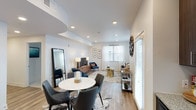 This single-floor residence features an open-concept layout that seamlessly connects the dining and living areas, creating a cohesive and functional living space. The interior is finished with light-toned hardwood-style flooring that runs throughout, contributing to a bright and airy atmosphere.

The dining area is anchored by a round marble-top table paired with charcoal upholstered chairs, positioned atop a cowhide-style area rug. A large floor-length mirror leans against the wall nearby, adding depth to the space.

The living area extends toward a windowed wall dressed with sheer white curtains and horizontal blinds that allow ample natural light to filter in. A coordinated seating arrangement featuring navy blue accent chairs surrounds a gold side table, complementing the neutral, warm tones of the space. A media console with a wall-mounted television anchors the living zone.

Recessed lighting is installed throughout the ceiling, supplemented by decorative table lamps, ensuring well-distributed illumination. The white walls and smooth ceilings maintain a clean, contemporary aesthetic throughout the open-plan space.