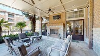 This elegant covered outdoor living space in Columbia, South Carolina, is designed around a commanding floor-to-ceiling fireplace constructed of warm brick and natural stone, serving as the focal point of the entire area. A wall-mounted flat-screen television above the fireplace makes the space equally suited for relaxed entertainment and social gatherings. The ceiling features a refined coffered design with recessed lighting and ceiling fans that provide airflow and comfort throughout the seasons. Large French doors with transom windows connect the covered patio to an interior space, maintaining an open and connected feel between indoors and out. The flooring combines large-format stone tiles with decorative gravel areas, adding texture and visual interest underfoot. Generously cushioned seating arrangements, including sofas, lounge chairs, and accent tables, are thoughtfully positioned to encourage conversation and relaxation around the fireplace. Ivy-draped stone columns frame open views to the surrounding landscaped grounds, which feature palm trees, manicured shrubs, and lush greenery. The overall ambiance blends classic Southern architectural charm with inviting outdoor comfort, creating a sophisticated gathering space that seamlessly integrates natural elements with stylish, functional design.