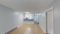 This single-floor space offers a clean, modern aesthetic with a bright and open layout that feels welcoming from the moment you step inside. The living area is finished with light hardwood-style flooring that extends seamlessly throughout, complementing the crisp white walls and white baseboard trim for a cohesive, airy feel. A baseboard heating unit runs along the wall, providing efficient warmth throughout the space.

The kitchen is positioned toward the rear of the living area and features white cabinetry, a dark countertop peninsula, and a window that invites natural light into the cooking space. The peninsula serves as a natural divider between the kitchen and the main living area, offering both functional prep space and a casual dining or gathering surface.

Beyond the kitchen, a short hallway leads to additional areas of the unit, with a door visible at the end suggesting access to a private entryway or additional room. The hallway features tile or patterned flooring, providing a practical transition zone. Flush-mount ceiling lighting ensures even illumination throughout. Overall, the space presents a well-maintained, functional layout with neutral finishes that offer a versatile foundation for a variety of living arrangements.