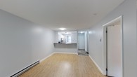 This single-floor space offers a clean, modern aesthetic with a bright and open layout that feels welcoming from the moment you step inside. The living area is finished with light hardwood-style flooring that extends seamlessly throughout, complementing the crisp white walls and white baseboard trim for a cohesive, airy feel. A baseboard heating unit runs along the wall, providing efficient warmth throughout the space.

The kitchen is positioned toward the rear of the living area and features white cabinetry, a dark countertop peninsula, and a window that invites natural light into the cooking space. The peninsula serves as a natural divider between the kitchen and the main living area, offering both functional prep space and a casual dining or gathering surface.

Beyond the kitchen, a short hallway leads to additional areas of the unit, with a door visible at the end suggesting access to a private entryway or additional room. The hallway features tile or patterned flooring, providing a practical transition zone. Flush-mount ceiling lighting ensures even illumination throughout. Overall, the space presents a well-maintained, functional layout with neutral finishes that offer a versatile foundation for a variety of living arrangements.