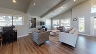 This single-floor space showcases a thoughtfully designed open-concept layout that seamlessly connects a comfortable living area with a generous dining space. The architecture is defined by striking vaulted ceilings with exposed wooden beams that lend the interior a sense of height and airiness, further emphasized by clerestory windows that draw natural light deep into the space.

The living area features durable hardwood-style flooring in a warm, medium-toned finish that extends throughout. Track lighting along the beamed ceiling provides flexible illumination, complementing the natural light that pours in through multiple large windows fitted with horizontal blinds. The living room accommodates a seating arrangement centered around a wall-mounted television and entertainment system, with a textured area rug anchoring the space.

A grand double-door entry with sidelights and transom windows creates a welcoming focal point, flanked by additional windows that enhance the bright, open atmosphere. The dining area offers ample room for a round table and chairs, positioned beneath a ceiling fan near French doors that provide views of the outdoors. A decorative sideboard with a large mirror adds an elegant finishing touch, completing this versatile and well-appointed space.