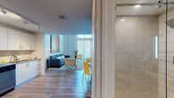 Located at 2160 Van Buren in Hollywood, Florida, this two-story apartment offers a sophisticated and contemporary living experience. The main level features an open-concept layout with light wood flooring that flows seamlessly throughout. The living area is anchored by soaring double-height ceilings and floor-to-ceiling windows fitted with vertical blinds, flooding the space with natural light. Bold artwork accents the walls, lending the space a curated, gallery-like ambiance. A glass-top dining table with white chairs sits beneath a striking black-and-white large-format art piece, creating a defined yet open dining area. The kitchen is outfitted with white shaker cabinetry, granite countertops, and a full suite of stainless steel appliances, including a refrigerator, range, microwave, and dishwasher, all set on warm wood-tone flooring. A clean, well-appointed bathroom on the main level features a granite vanity, illuminated mirror, and a glass-enclosed shower with tile surround. The upper level serves as a private retreat, housing the spacious primary bedroom with access to an en-suite bathroom, a dedicated workspace, and an open loft-style overlook with sleek black metal railings and expansive windows offering outdoor views.