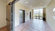 This single-floor space showcases a functional yet thoughtfully designed interior typical of a multi-story commercial or residential building. The elevator lobby serves as the central hub of the floor, housing two stainless steel elevator doors framed by light-colored walls. A floor-to-ceiling window wall floods the lobby with natural light and offers views of the surrounding outdoor landscape, including trees and neighboring structures.

The corridors and common areas feature durable terracotta-toned tile flooring that runs consistently throughout, complementing the warm yellow-toned painted walls. Recessed ceiling lighting provides even, ambient illumination across the spaces. Decorative framed photographs depicting sunset and coastal scenes are displayed along the corridor walls, adding a welcoming, artistic touch to the otherwise utilitarian environment.

The floor is clearly marked with a large numeral "3," identifying it within the building. Stairwell access is provided via a solid gray fire door, with exit signage prominently displayed at key transition points to ensure clear wayfinding and safety compliance. The overall ambiance is clean, bright, and straightforward, balancing practical building infrastructure with modest aesthetic details that contribute to a comfortable and navigable common area.