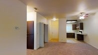 This single-floor residence offers a straightforward and functional layout designed for comfortable everyday living. The main living area is generously sized, finished with wall-to-wall carpeting that adds warmth underfoot. A ceiling fan with an integrated light fixture is centrally mounted, providing both air circulation and ambient lighting. A large sliding glass door floods the space with natural light and opens to an outdoor area, framed by vertical blinds for privacy and light control. The walls are painted in a neutral, warm tone that complements the soft carpeting throughout.

Adjacent to the living space, a transitional hallway area connects to additional rooms, with a bi-fold closet door and a dark wood interior door visible, suggesting well-organized storage solutions. The flooring transitions from carpet to a hard surface in this zone, adding practical durability near entryways.

The kitchen is positioned to one side and features dark cabinetry contrasted against lighter countertops, a built-in dishwasher, and overhead fluorescent lighting that ensures a bright workspace. A window above the sink introduces natural light into the kitchen area. The overall layout is efficient, with all essential areas thoughtfully connected across a single level.