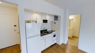 This single-floor unit offers a clean, functional layout highlighted by a well-organized kitchen alcove. The kitchen is fitted with white cabinetry both above and below the counter, providing ample storage. Essential appliances include a full-size refrigerator, an electric range, a microwave, and a sink with a dark countertop — all presented in a cohesive white finish that contributes to a bright, tidy aesthetic. The kitchen is neatly recessed into a dedicated nook framed by white walls, giving it a structured, built-in appearance.

Warm hardwood flooring runs through the main living area, adding a natural contrast to the predominantly white interior. The open sightlines from the kitchen toward adjoining spaces suggest a straightforward, efficient floor plan. A glimpse into an adjacent area reveals light tile flooring and additional cabinetry, likely a bathroom or utility space. The overall ambiance is minimal and clean, with a neutral color palette that creates a bright, open feel throughout the unit.