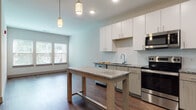 This contemporary single-floor unit offers a thoughtfully designed open-concept layout that seamlessly connects the kitchen and living area. The kitchen is a standout feature, equipped with stainless steel appliances including a range, microwave, dishwasher, and refrigerator. White flat-front cabinetry provides ample storage, while granite countertops extend across the workspace and onto a central island that doubles as a dining and prep surface. A classic white tile backsplash, pendant lighting overhead, and warm wood-look flooring throughout contribute to the space's clean, modern aesthetic. Natural light filters into the living area through large windows, creating a bright and open atmosphere. A dedicated wine rack cabinet adds a charming and functional touch near the refrigerator. The bathroom is finished with the same attention to detail, featuring a full soaking tub with white subway tile surround, a granite-topped vanity with modern hardware, and wood-look flooring for a cohesive look. The bedroom offers a comfortable retreat with plush wall-to-wall carpeting and neutral tones throughout. Together, the rooms present a well-balanced, move-in-ready space with consistent modern finishes and a clean, inviting ambiance.