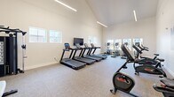 Located at 2507 Orogrande Ln in Caldwell, Idaho, this dedicated fitness center offers a bright, open workout environment designed to support a range of exercise routines. The space is defined by its striking vaulted ceiling, which rises dramatically to create an airy, expansive atmosphere that sets it apart from a typical gym setting. Suspended linear LED lighting runs along the roofline, providing consistent, even illumination throughout the room.

The interior is finished with neutral, light-toned walls and durable carpet flooring, contributing to a clean and modern aesthetic. A row of multiple treadmills lines one wall, accompanied by a wall-mounted flat-screen television, making cardio sessions both convenient and engaging. Adjacent to the treadmills, a bank of elliptical trainers is positioned in front of a series of windows that frame views of the surrounding community, allowing natural light to flood the space. Upright and recumbent stationary bikes are distributed throughout the room, offering low-impact cardiovascular options. A cable strength machine and a workout bench provide opportunities for resistance training as well.

Fitness-themed artwork decorates one wall, adding a motivational touch to the space. A dedicated entry door and visible safety equipment reflect thoughtful facility planning. Overall, this fitness center delivers a well-rounded, welcoming environment for residents seeking convenient, on-site exercise amenities.